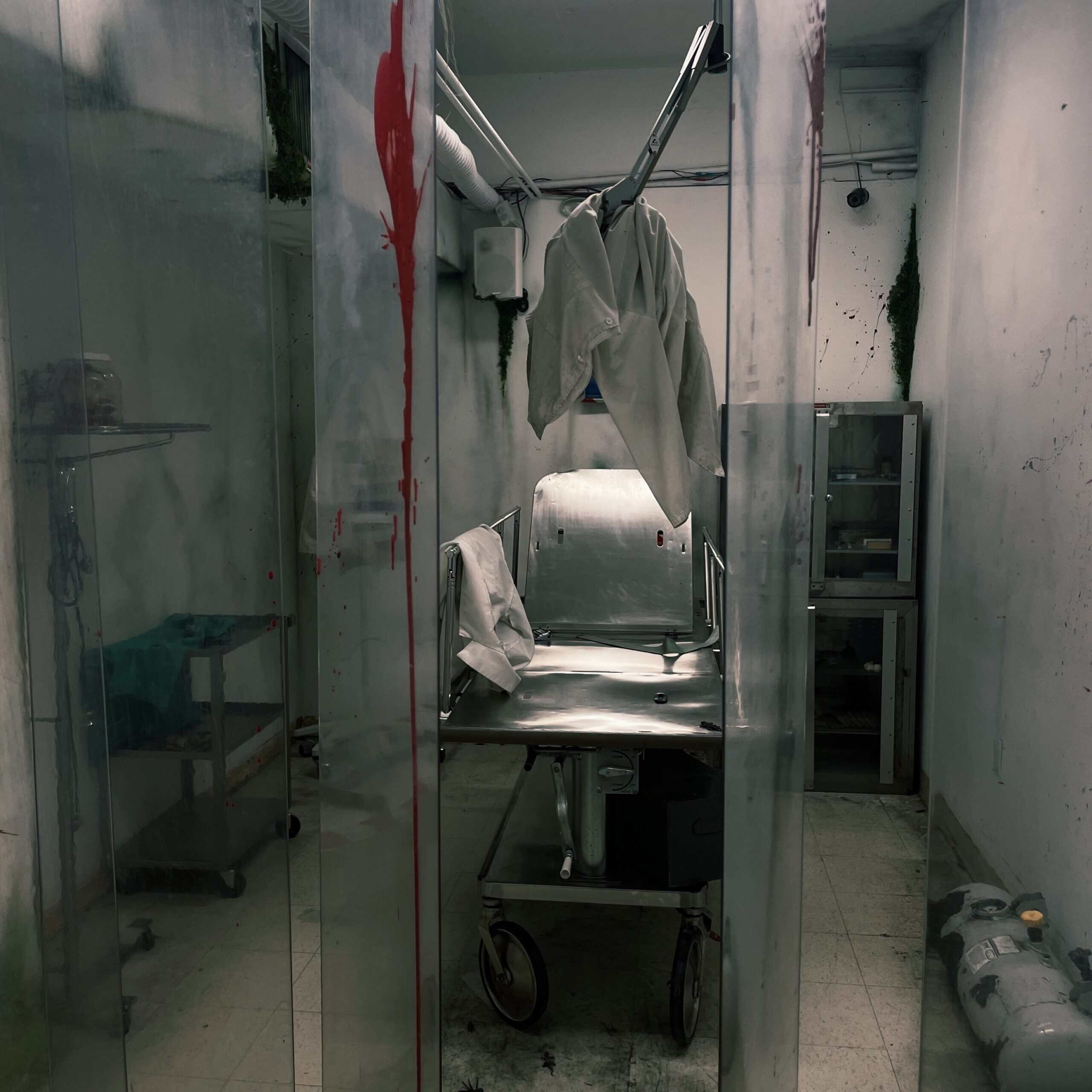 Blood-stained hospital gurney in a dark, abandoned medical room with hanging clothes.