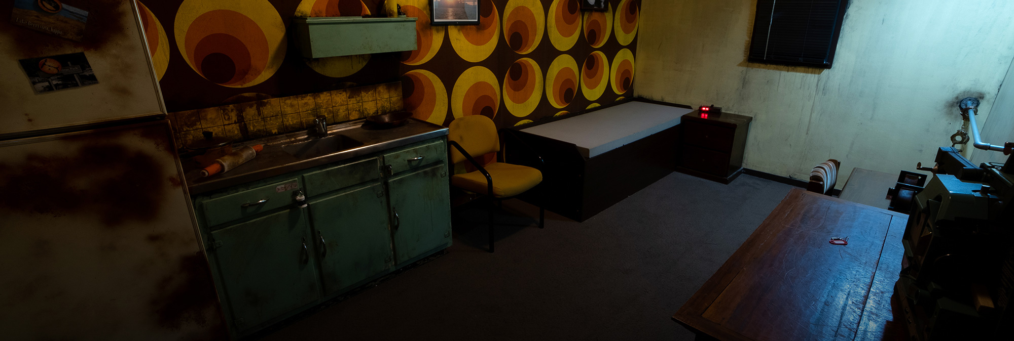 Faded kitchen with retro green cabinets, yellow patterned wall, and dim lighting in escape room.