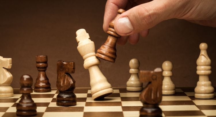 Dark brown and light beige chess pieces on a wooden chessboard, hand moving a white queen piece, strategic game scene, Roomescape by Fox in a Box Stockholm.
